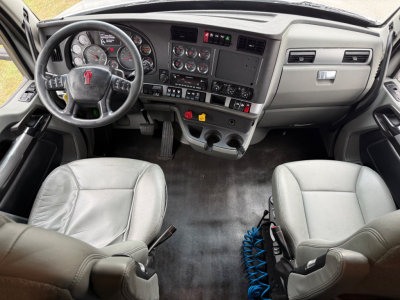 Truck cab interior showing steering wheel with Kenworth logo, dashboard with gauges, center console with controls, two gray seats, floor mat, and blue coiled air hose.