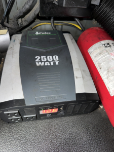 In-vehicle view of a 2500-watt Coburn? inverter labeled 2500 WATT and 12V; visible control panel with gauge; red fire extinguisher beside; wiring and components in the engine bay area.