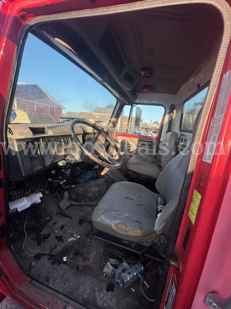Interior of a red 1999 International 4700 low profile truck cab showing worn driver's seat and dashboard, with visible steering wheel and pedals.