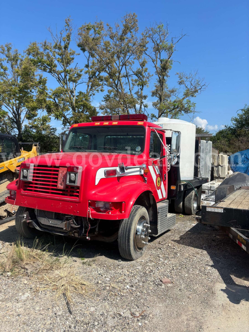 International 4700 low profile truck in red with white cab sleeper, front grille, dual headlights, flatbed with equipment in yard.