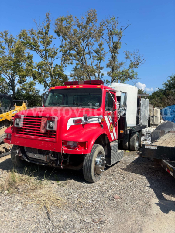 International 4700 low profile truck in red with white cab sleeper, front grille, dual headlights, flatbed with equipment in yard.
