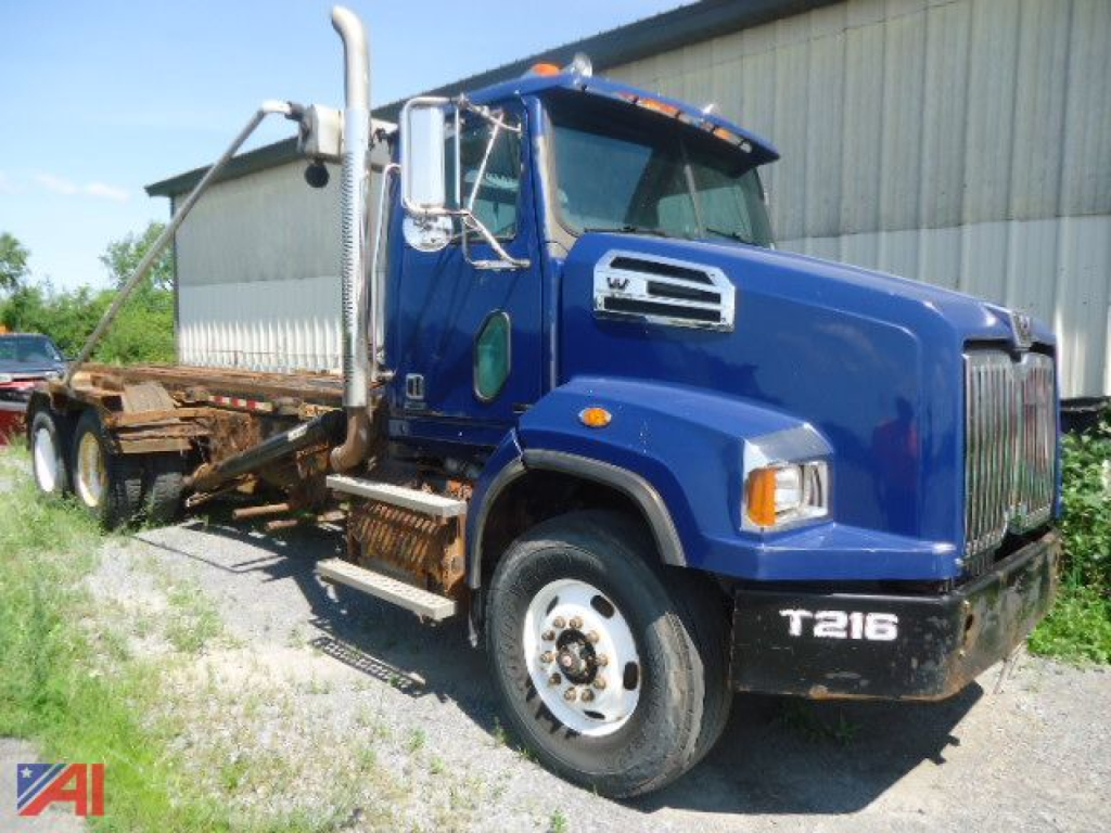 Blue 2015 Western Star 4700 SB Roll Off Truck with a flatbed and undercarriage visible, front grille, side exhaust, and T216 label on the bumper.