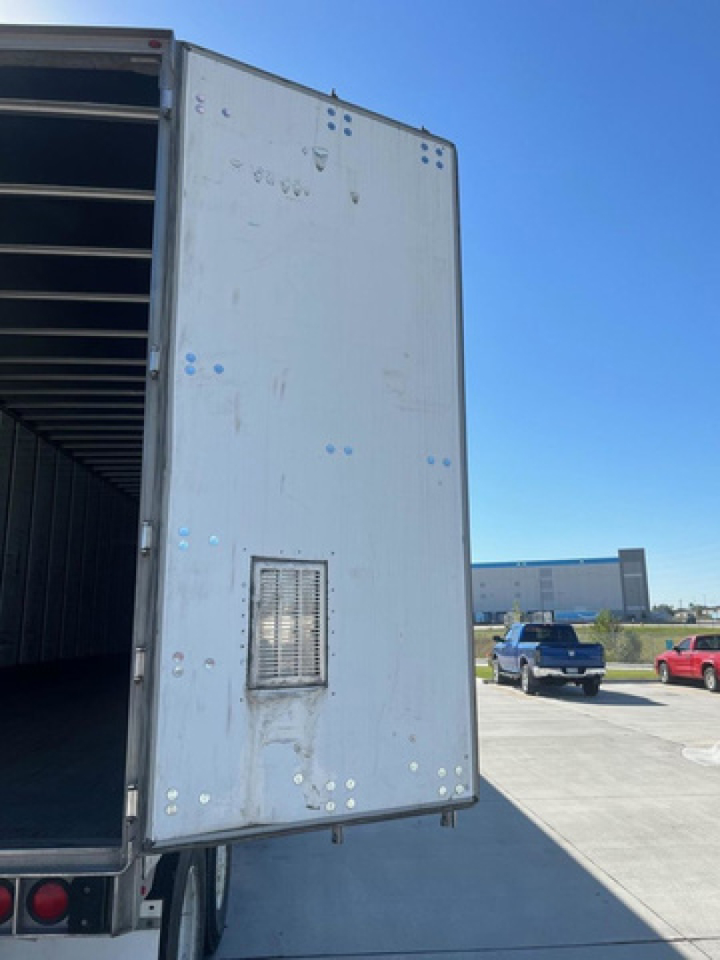 Truck trailer rear door open showing vertical metal panel with small vent and riveted edges; blue and white faded paint; clear sky and surrounding lot with vehicles in background.
