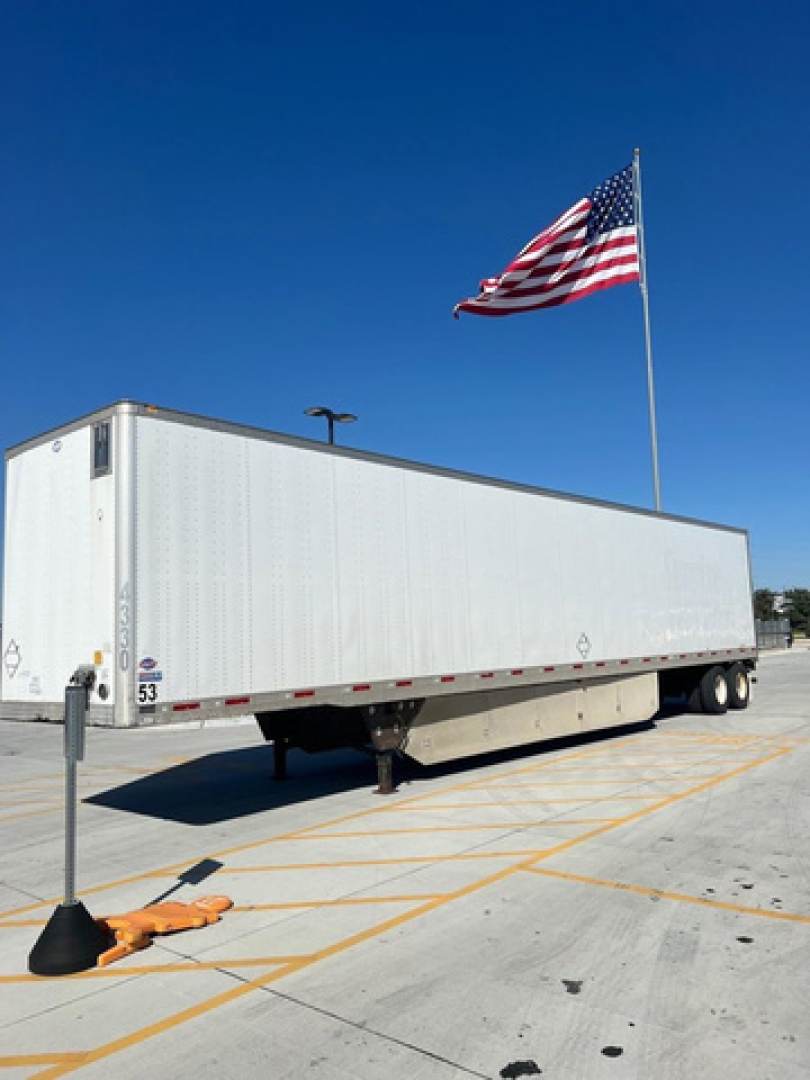 White utility trailer/van trailer with a long rectangular cargo area on a parking lot, American flag in background, under bright blue sky.