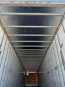 Trailer interior showing metal walls and ceiling ribs, creating a long narrow cargo space with a wooden floor at the far end.