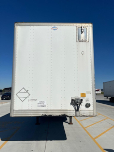 Rear view of a white utility trailer/van showing bumper area with hitch, electrical connections, warning decals, and a small vent door.