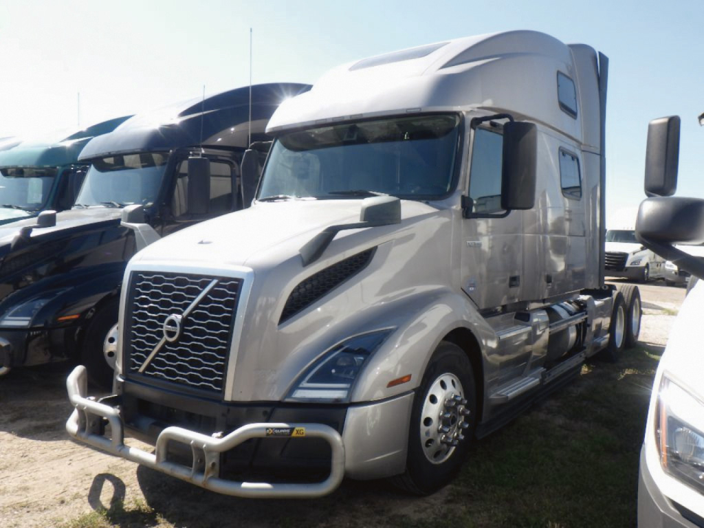 Truck showing 2023 Volvo VNL 860 with high sleeper, chrome grille, and bumper guards; front view in a lot with other trucks in background.