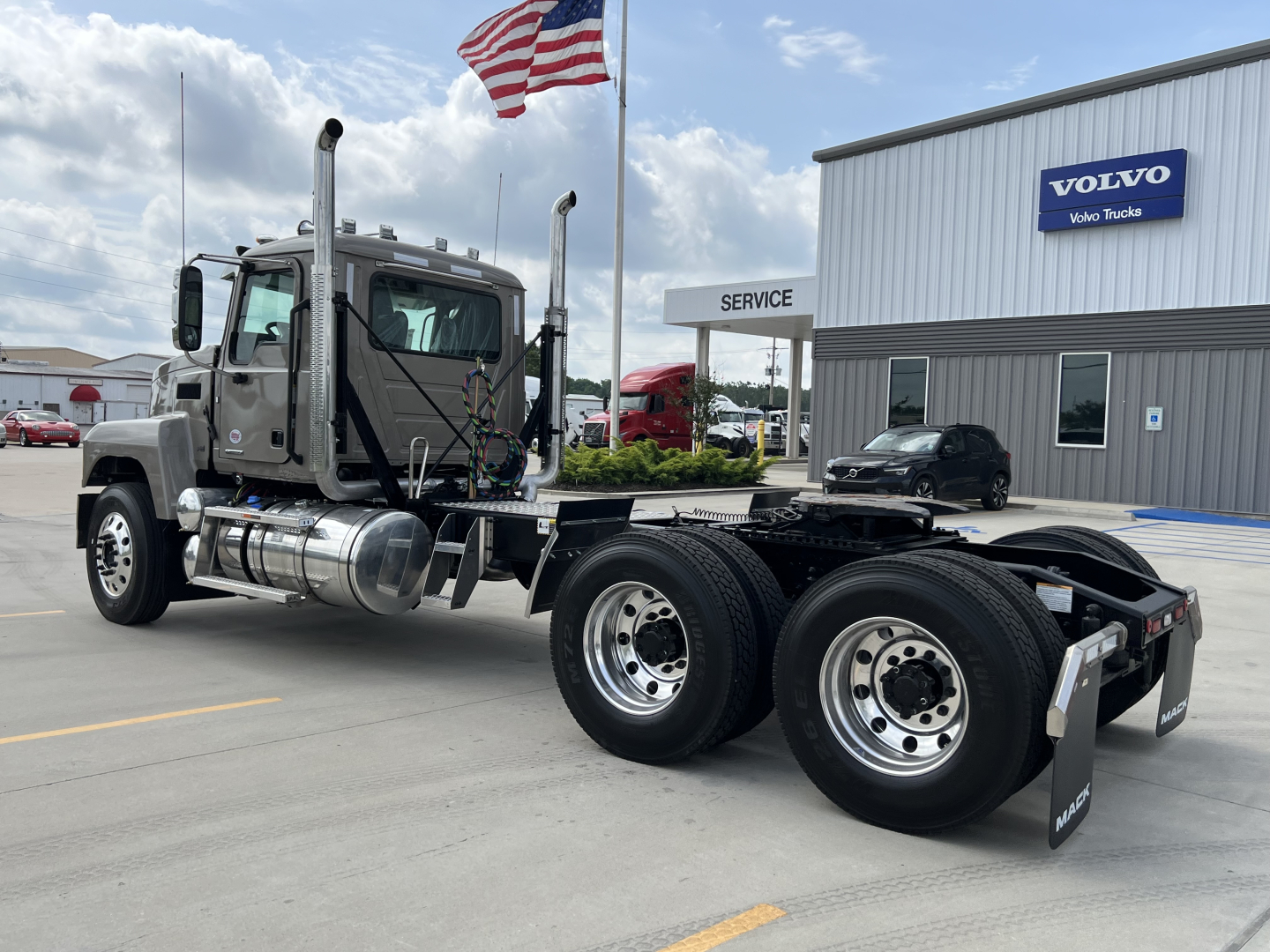 Truck chassis and cab of a 2027 Mack PI64T conventional daycab, dual rear axles, shiny wheels, fuel tanks, exhaust stacks, and a Volvo dealer building in background.