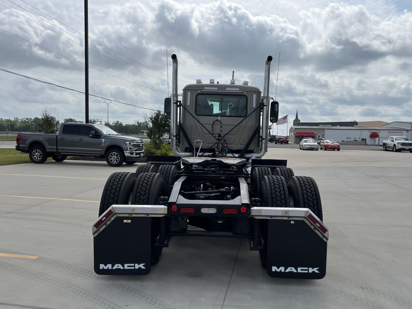 Rear view of a new 2027 Mack PI64T daycab tractor showing dual rear axles, mud flaps with 'MACK' branding, and chassis wiring harness.