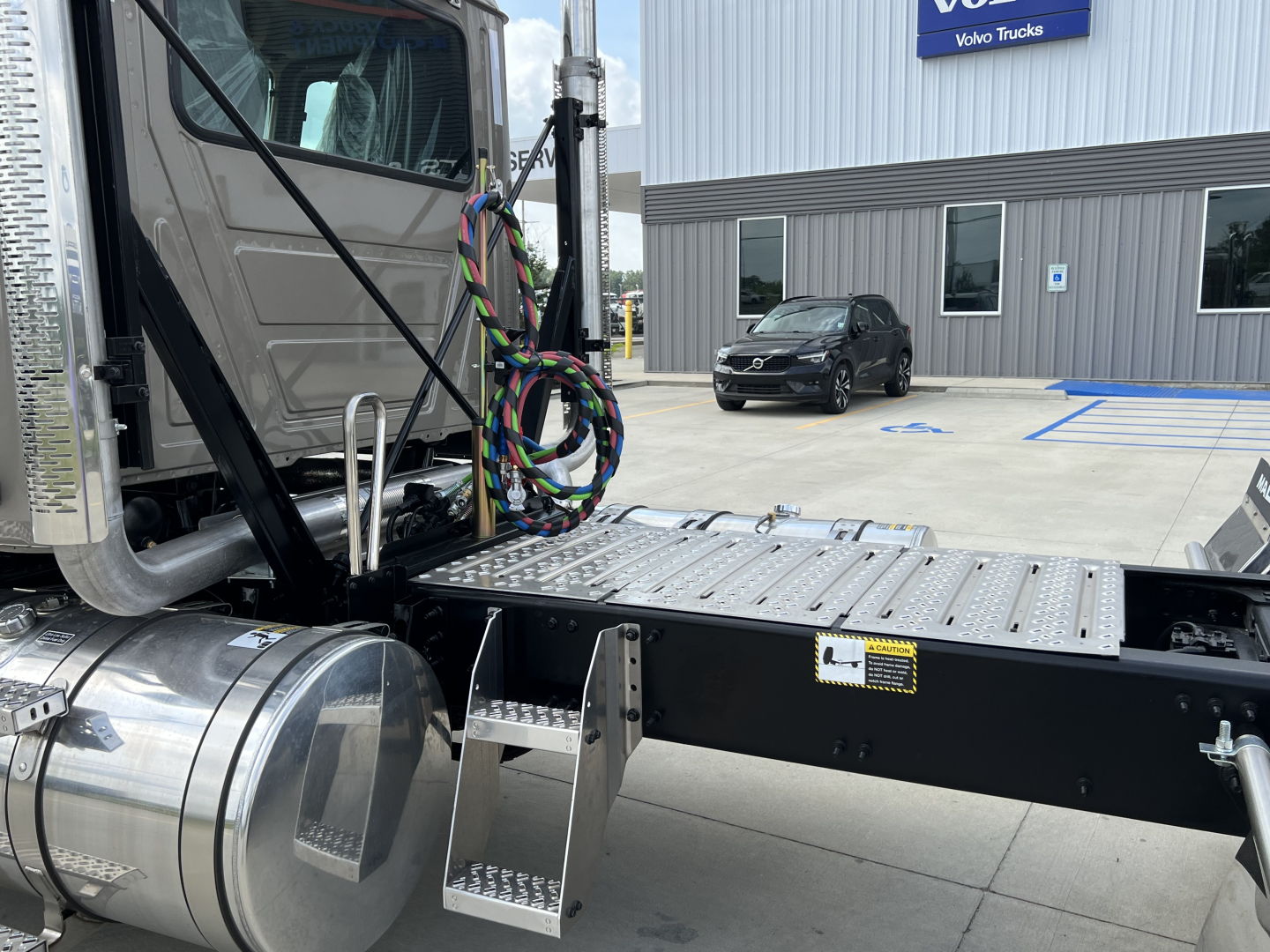 Side view of a Mack PI64T cab and chassis showing a metal step and chassis rails with wiring harnesses near the cab; other vehicles and a dealership building visible in background.