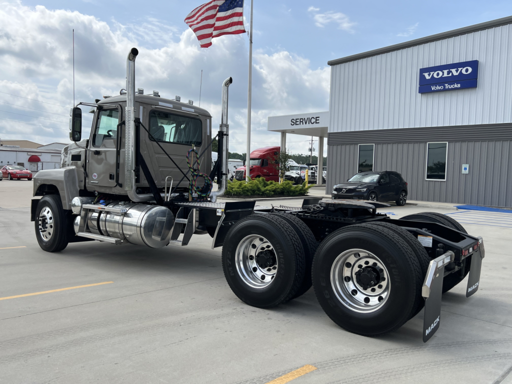 Truck chassis and cab of a 2027 Mack PI64T conventional daycab, dual rear axles, shiny wheels, fuel tanks, exhaust stacks, and a Volvo dealer building in background.