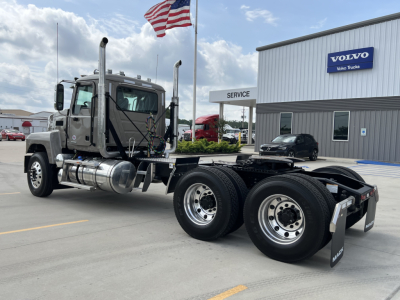 Truck chassis and cab of a 2027 Mack PI64T conventional daycab, dual rear axles, shiny wheels, fuel tanks, exhaust stacks, and a Volvo dealer building in background.