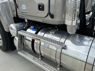 Tank and side view of a new Mack PI64T daycab with chrome fuel tanks, metal steps, and intake/exhaust system. Visible are the cylindrical aluminum fuel tank, access platform, blue cap, hoses, and chassis components.