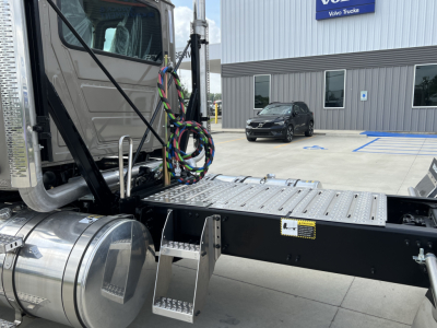 Side view of a Mack PI64T cab and chassis showing a metal step and chassis rails with wiring harnesses near the cab; other vehicles and a dealership building visible in background.