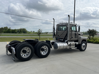 Mack PI64T conventional daycab tractor in showroom lot showing side profile with dual rear axles, fuel tanks, and exhaust stacks.