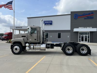 2027 Mack PI64T truck chassis in a dealership lot, gray cab, three rear axles, black chassis, fuel tanks under cab, dealership building in background with Volvo sign and American flag.
