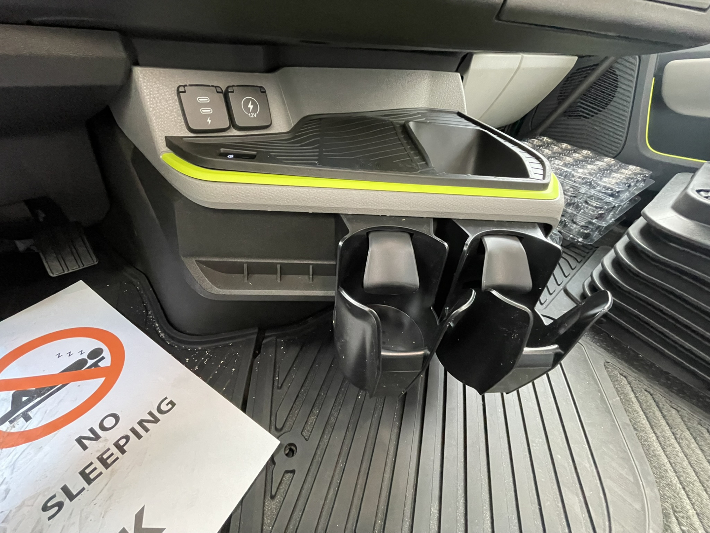 Inside cab area of a Volvo VNL truck showing a control panel, lime accent trim, and two cup holder slots near the floor mat.
