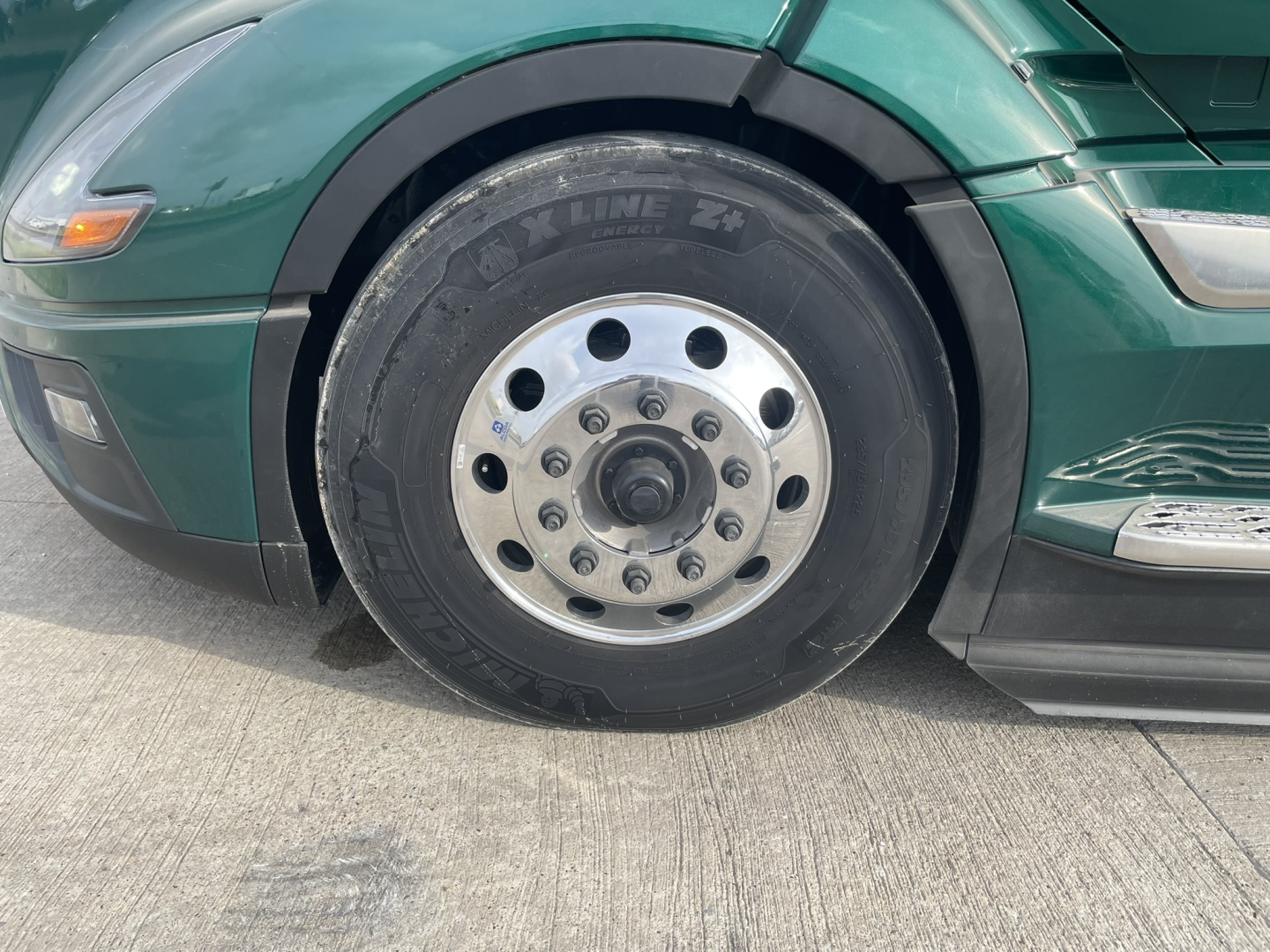 Truck wheel and tire close-up showing front front axle area of a green Volvo VNL64T860 sleeper cab with steel wheel and tire readout.