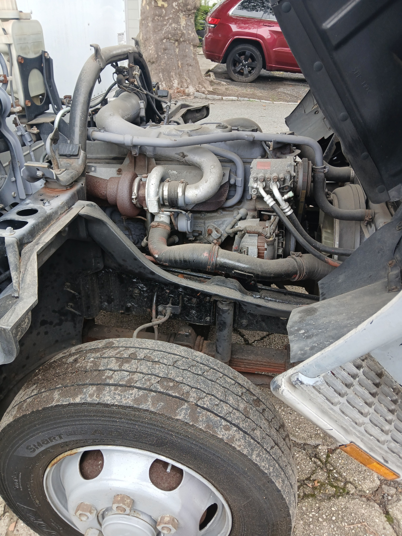Engine bay view of a 2004 Mitsubishi FH truck showing turbocharger piping, hoses, and engine components near front wheel.