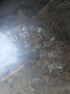 Gauge cluster close-up showing speedometer with numbers and a sediment of dirt; markings include 0-50-60 and 100, with a needle near 0; dirt obscures surrounding text.