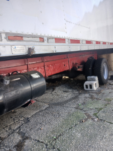Trailer undercarriage side view showing fuel tank and dual wheels with support block and chocks; white trailer wall with riveted panels.