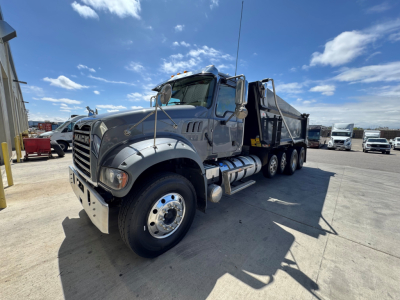 Dump truck showing Mack GR64F front-left angle with dual rear axles and dump bed. Visible text includes Mack grille and large chrome bumper.