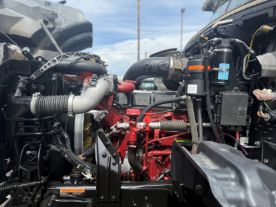 Dump truck engine bay of a 2024 Mack GR64F showing red engine block with hoses, cables, and components including air intake and fluid reservoirs.