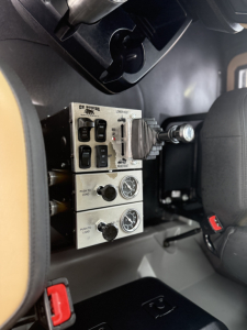 Truck interior showing control panels with switches and gauges mounted on a metallic panel near the driver seat.