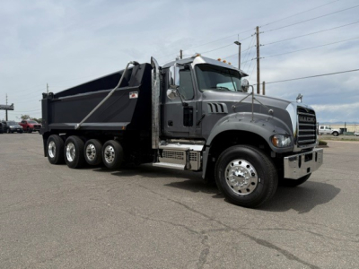 Dump truck in a parking lot with a gray Mack GR64F dump bed, side view showing six wheels on tandem rear, chrome bumper, and side steps.