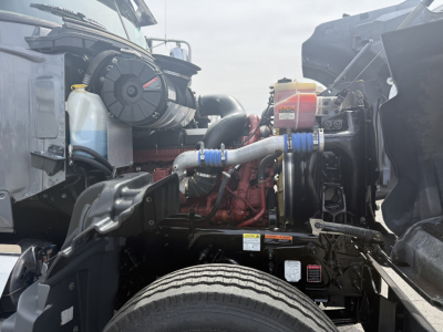 Mack GR64F engine bay showing coolant reservoir, air filter canister, turbocharger pipework, and radiator hoses.