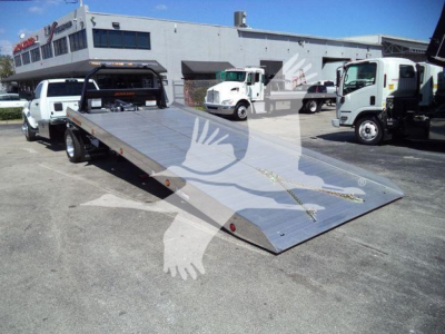 A flatbed tow truck with a metallic silver ramp in the foreground and several white trucks in the lot behind, dealership building in background.