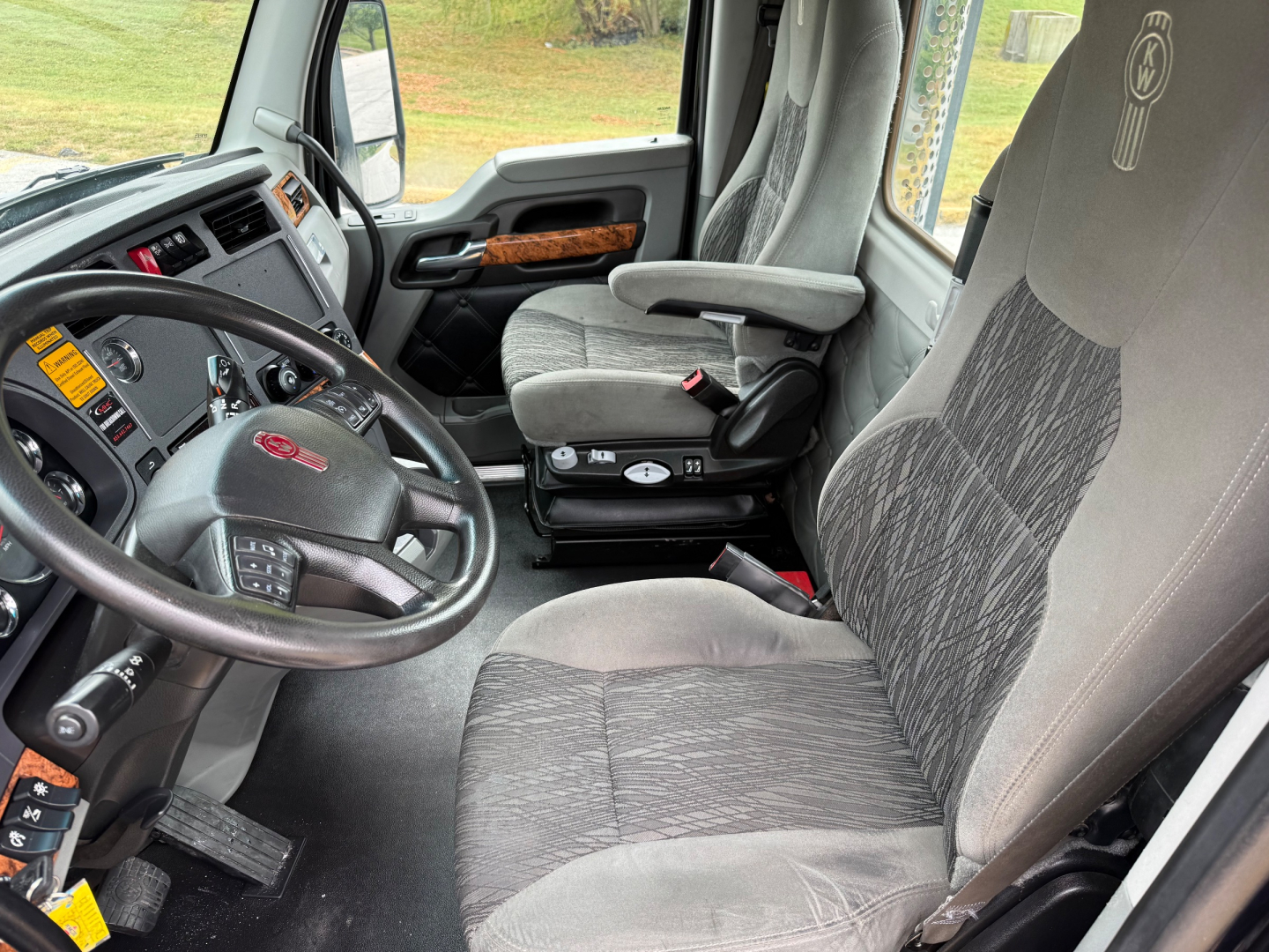 Kenworth T880 truck interior showing steering wheel, dashboard with controls, driver seat, and missing gear shifter area. Seats are fabric with gray and black pattern.