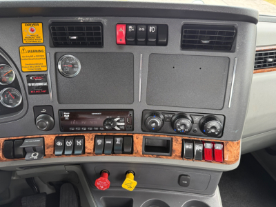 Kenworth T880 dash panel with warning stickers, gauges, controls, and woodgrain trim; features yellow and red hazard switches, air vents, and climate controls.