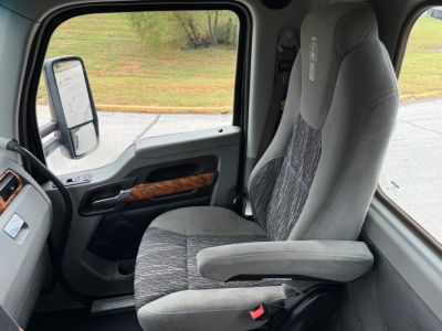 Kenworth T880 interior driver seat and dashboard as seen through the cab door, grey fabric seat with armrest, woodgrain trim on door, side mirror visible.