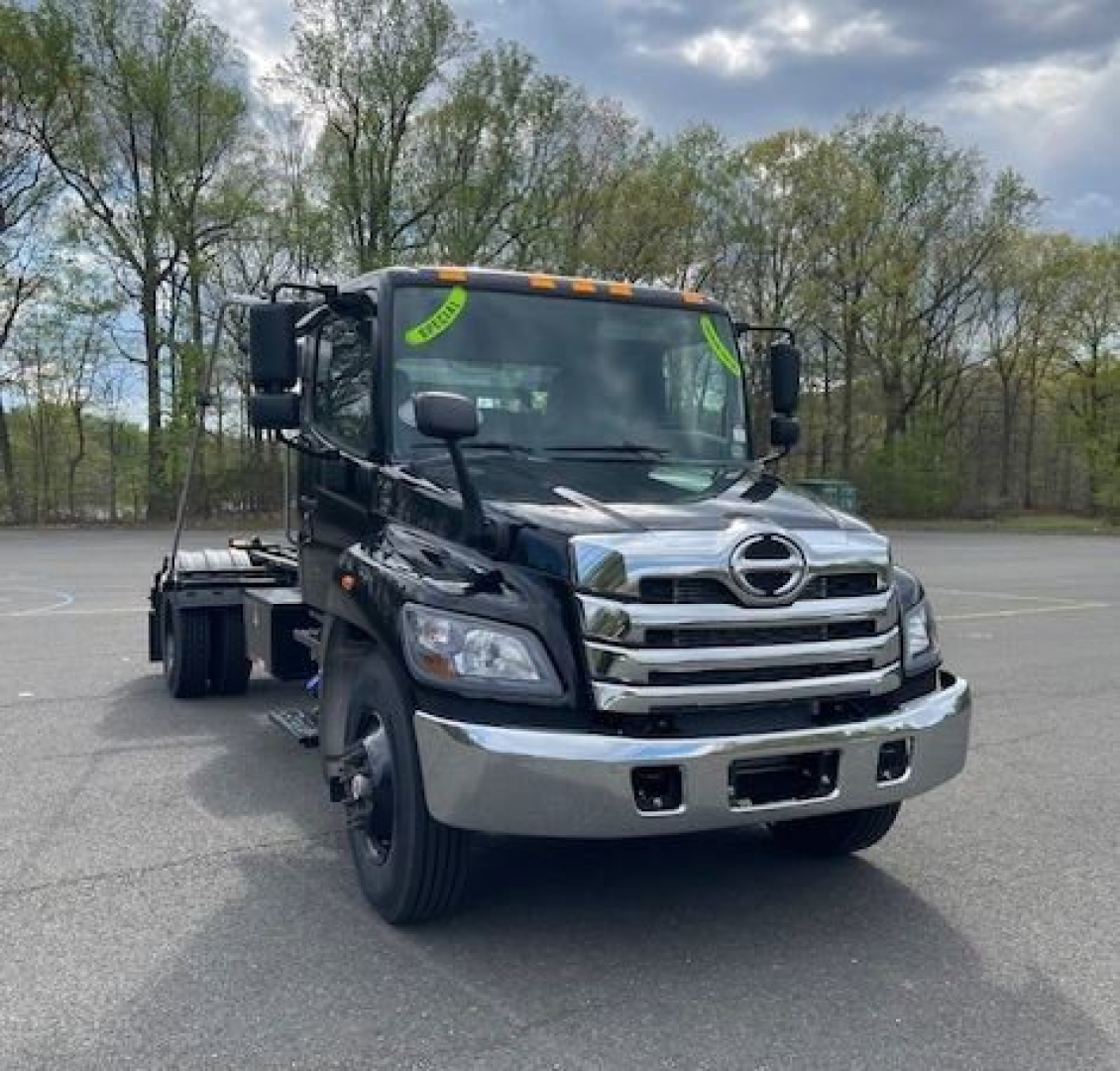 Front view of a black 2027 Hino L7 hooklift truck chassis with chrome grille and bumper, single front wheel visible, green inspection stickers on windshield.