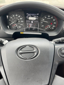 Dashboard of a Hino truck with steering wheel centered, showing instrument cluster and digital readout. Visible text includes Hino logo on steering wheel, numbers on gauges, gear indicator showing N, and central display with faint text.