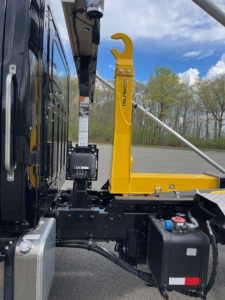 Truck side view showing yellow Paulinager hooklift arm and black cab chassis with hydraulic components and battery box.