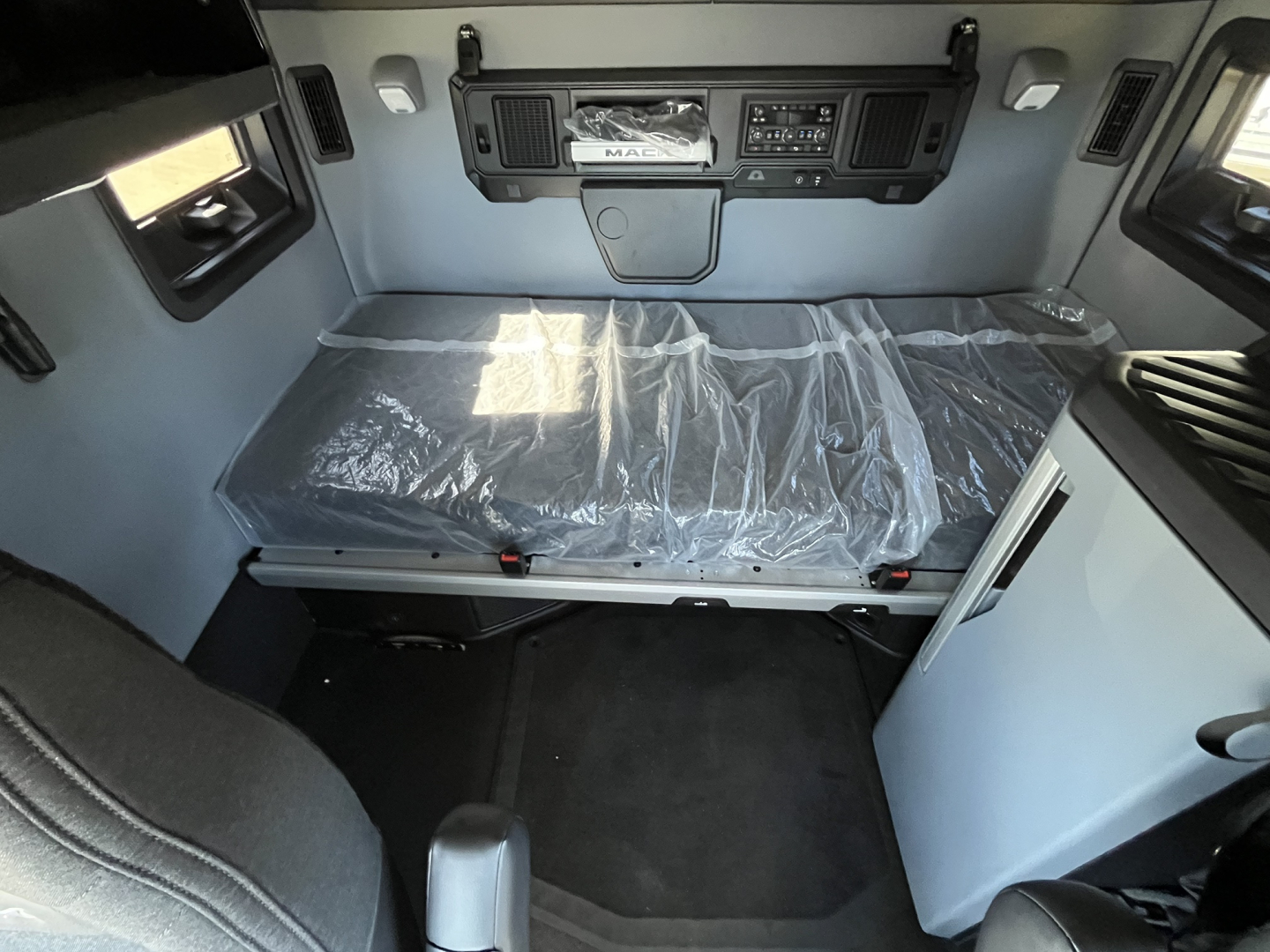 Mack PR64T76M truck cab interior showing sleeper berth with plastic-covered mattress, dashboard controls, and side storage panels.