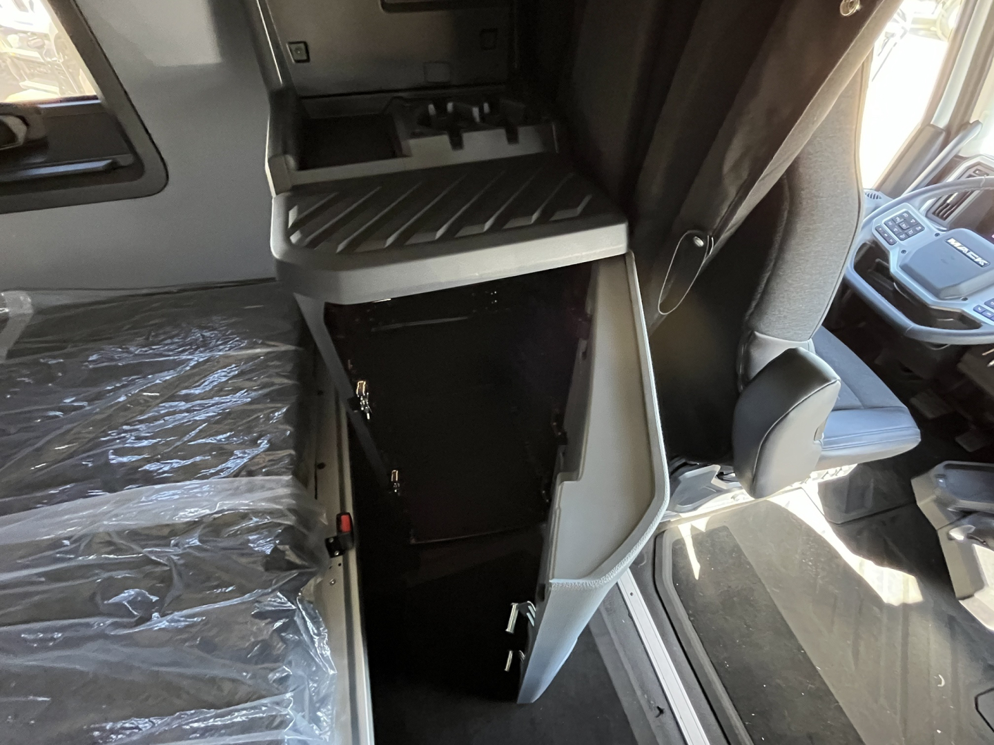 Cab interior of a new Mack PR64T76M showing a storage compartment door open, with seat plastic covering and dash area visible.