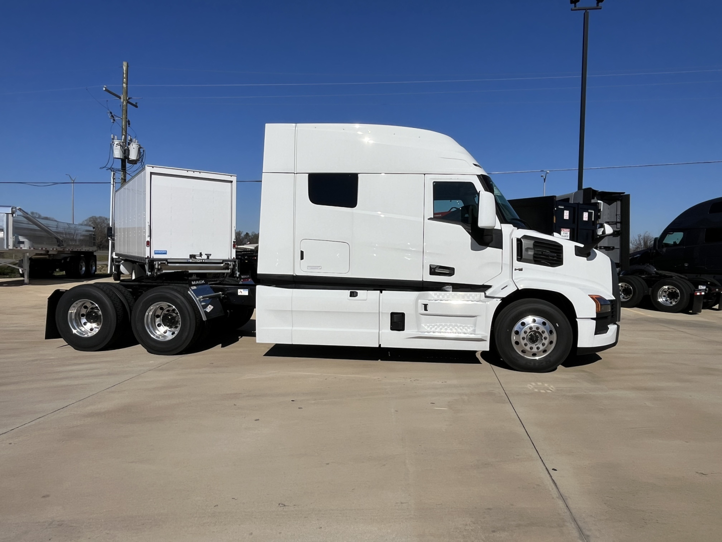 Truck side view showing 2026 Mack PR64T76M conventional sleeper with white exterior and dual rear axles, cab profile, and attached trailer in a lot.