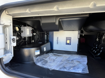 Mack PR64T76M sleeper truck engine bay showing auxiliary components and a white Xantrex box; plastic bag on floor mat; various wiring and black cylindrical reservoir visible.