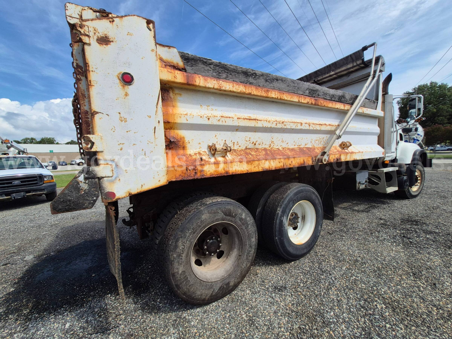 Dump truck rear/side view showing a white Mack CV713 dump bed with rust along edges, dual rear axle, and hydraulic tailgate. Visible text includes stock vehicle signs and general truck components.