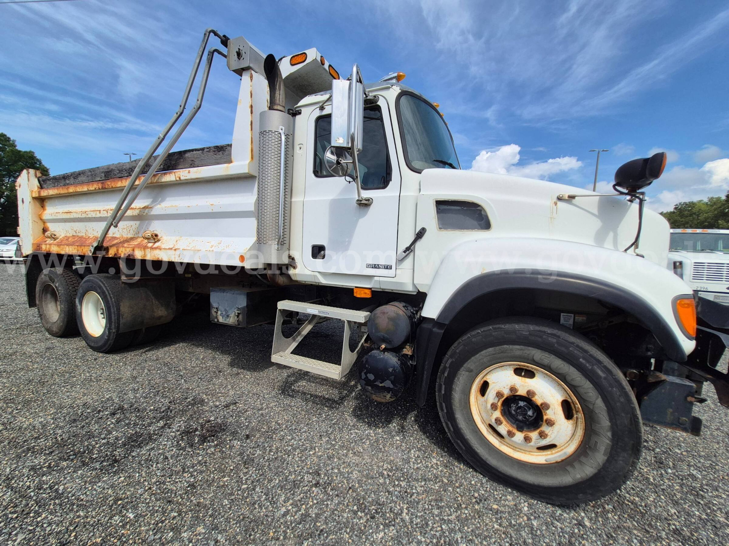 Dump truck side view showing cargo bed and Mack CV713 badge with white cab and rust on wheel rims.