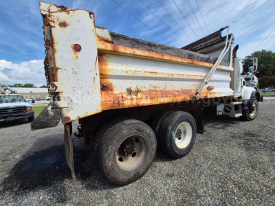 Dump truck rear/side view showing a white Mack CV713 dump bed with rust along edges, dual rear axle, and hydraulic tailgate. Visible text includes stock vehicle signs and general truck components.