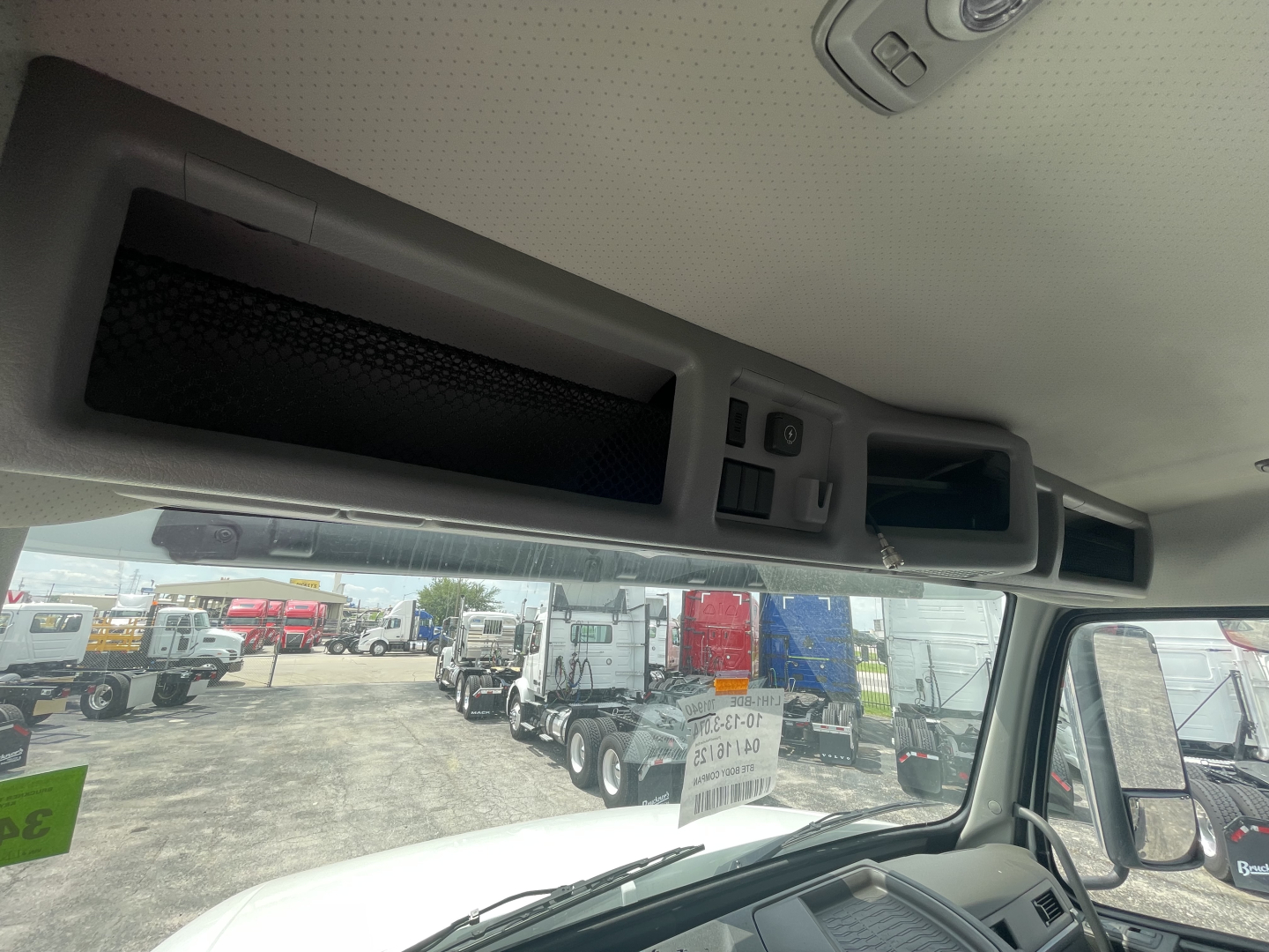 Interior overhead control panel and storage compartments in a Volvo VHD truck cab. Visible are ceiling grab handles, switches, and vents; through the windshield a truck yard with multiple red and blue trailers.
