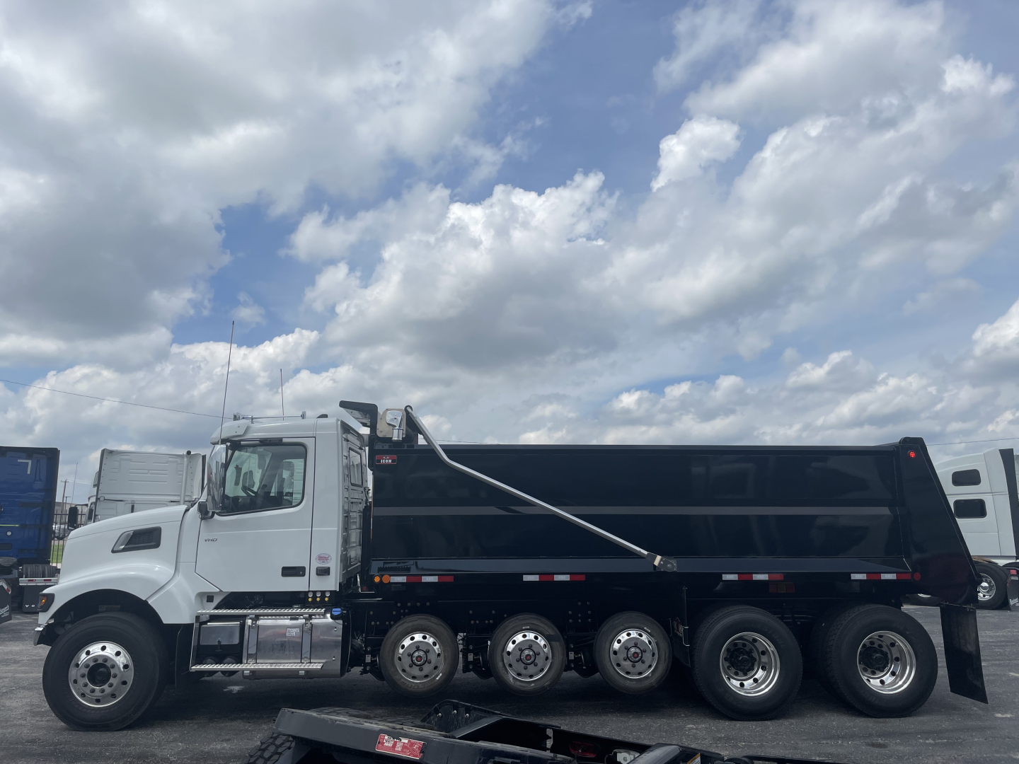 Dump truck shown: 2026 Volvo VHD64F300 in white cab with black dump body, multiple axles (six wheels on rear), daytime sky with clouds.