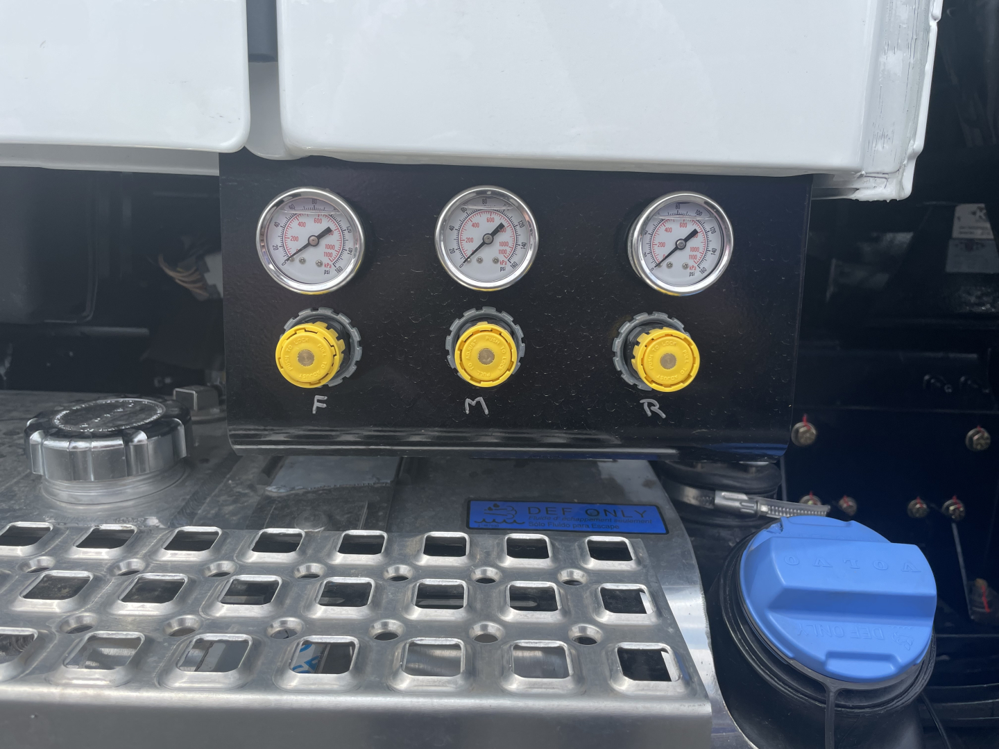 Volvo VHD64F300 engine bay panel with three pressure gauges and yellow knobs labeled F M R, blue coolant cap, metal step grid.