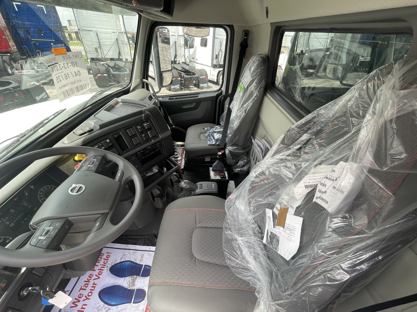 Truck interior: Driver seat and passenger seat covered in plastic; steering wheel; dashboard with controls; center console; large side window; steering column; plastic-wrapped seats and tags.