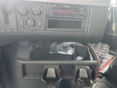 Dashboard area of a Volvo VHD64F300 truck interior showing climate controls, aftermarket stereo, and open storage compartments with a plastic bag and assorted items.