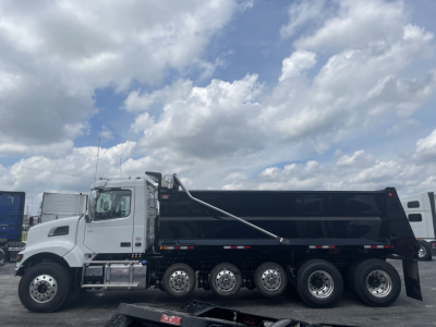Dump truck shown: 2026 Volvo VHD64F300 in white cab with black dump body, multiple axles (six wheels on rear), daytime sky with clouds.